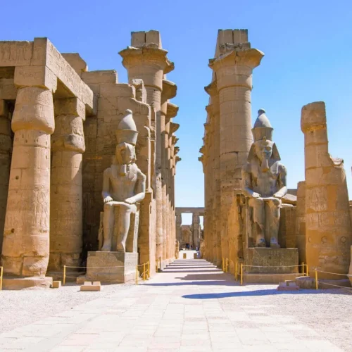 seated granite royal statues of ramesses ii flanking the entrance pylon of luxor temple under blue sky