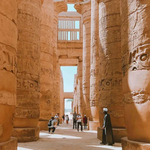 travelers walking among towering carved sandstone pillars inside the great hypostyle hall of karnak temple