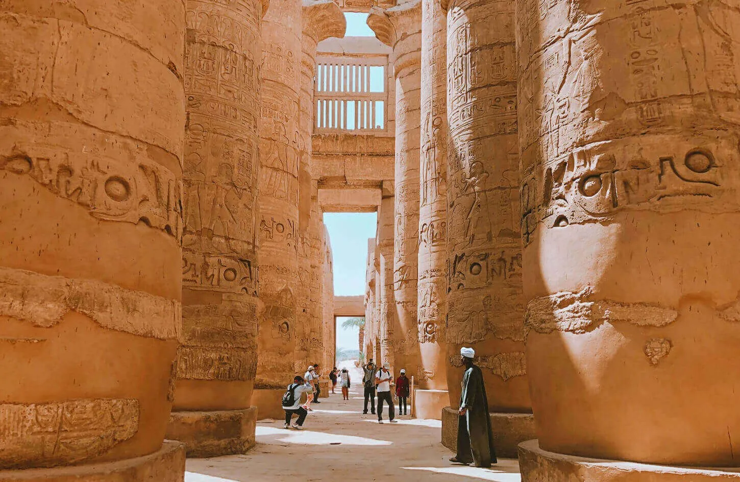 travelers walking among towering carved sandstone pillars inside the great hypostyle hall of karnak temple