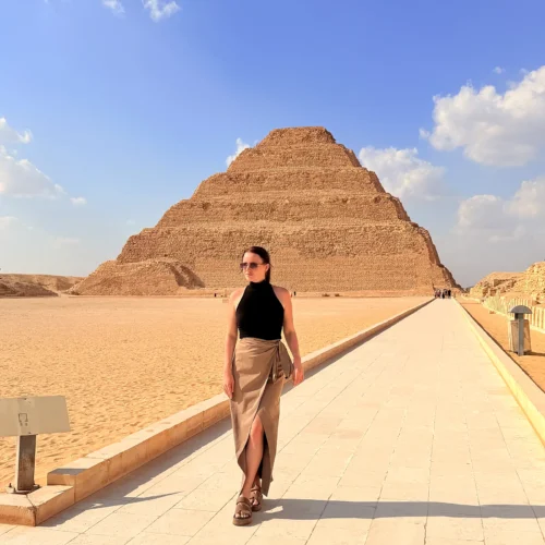 Traveler in sunglasses walking along a paved path with the Step Pyramid of Djoser rising behind her at Saqqara