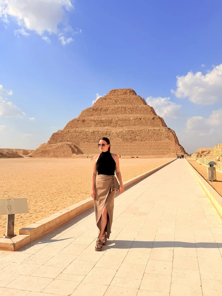 Traveler in sunglasses walking along a paved path with the Step Pyramid of Djoser rising behind her at Saqqara