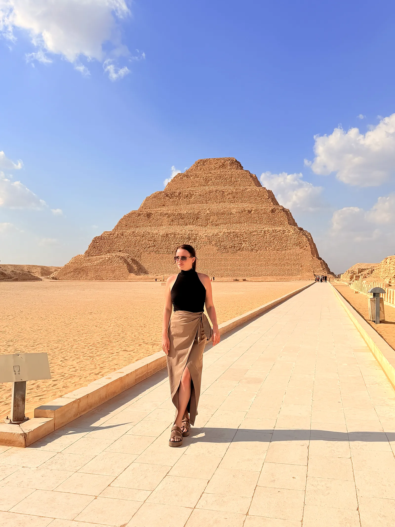 Traveler in sunglasses walking along a paved path with the Step Pyramid of Djoser rising behind her at Saqqara