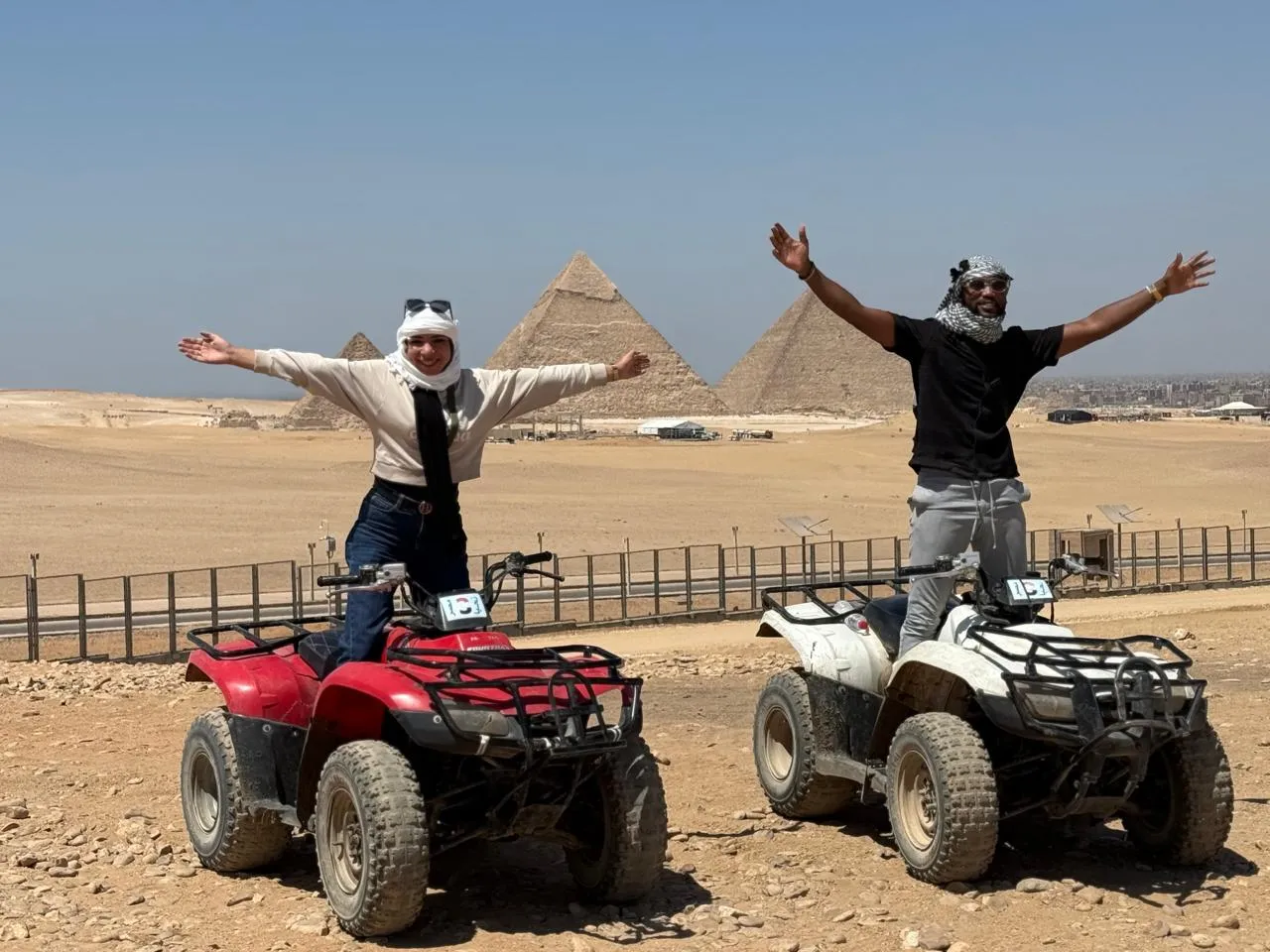 Couple standing on red and white ATVs with arms raised before the Giza pyramids