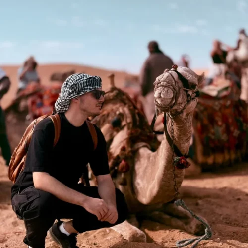 Traveler in a keffiyeh crouching beside a seated camel in the Giza desert