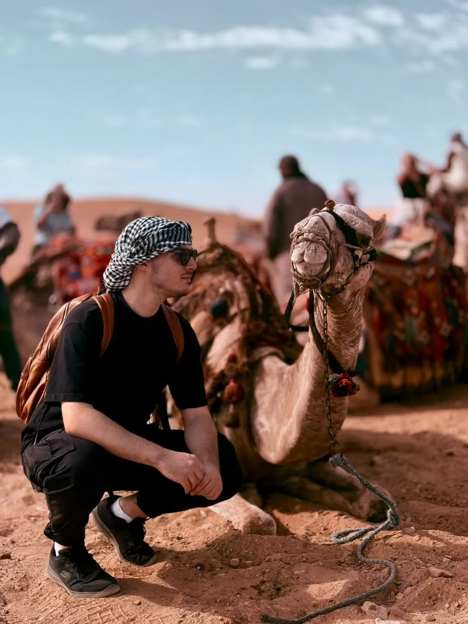 Traveler in a keffiyeh crouching beside a seated camel in the Giza desert