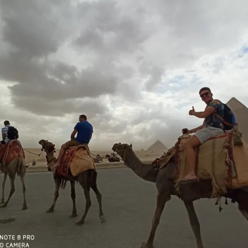 Travelers riding camels across the Giza Plateau with pyramids behind