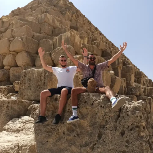 Two travelers seated with arms raised on the blocks of a Giza pyramid
