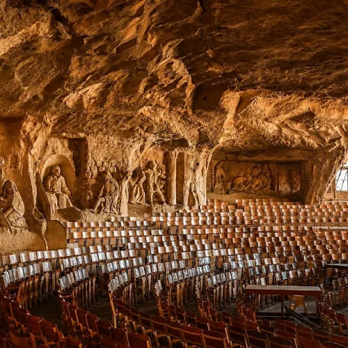 Interior of St. Simon Cave Church amphitheater with wooden chairs and large biblical bas-reliefs carved into the rock walls