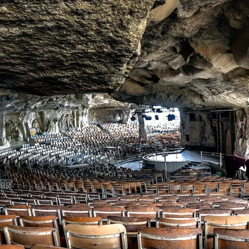 Vast rock-cut amphitheater church at St. Simon monastery with tiered wooden chairs facing a circular stage under the cave ceiling