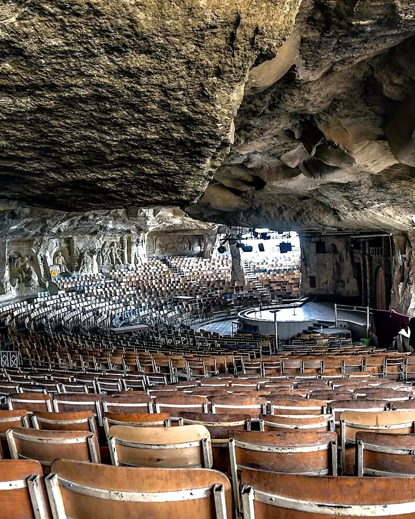 Vast rock-cut amphitheater church at St. Simon monastery with tiered wooden chairs facing a circular stage under the cave ceiling