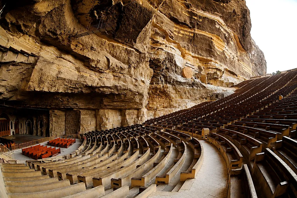 Open amphitheater seating under stratified Mokattam cliff rock with carved walls at St. Simon Cave Church