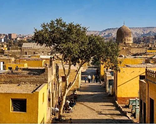 Sandy street in Cairo’s City of the Dead lined with yellow buildings, a central tree, and stone domes beyond