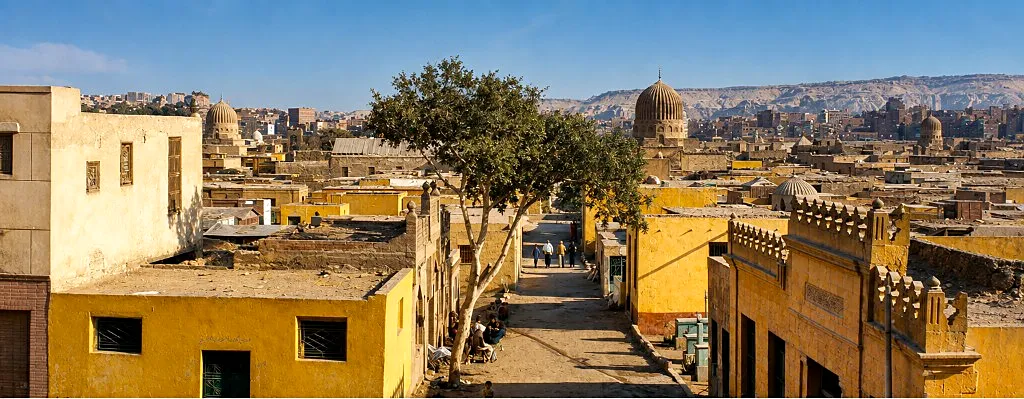 Sandy street in Cairo’s City of the Dead lined with yellow buildings, a central tree, and stone domes beyond