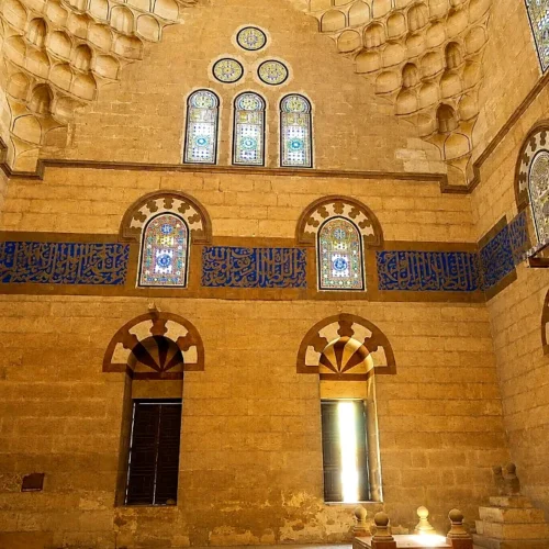 Interior of a Mamluk funerary mosque with blue calligraphy band, stained-glass windows, and stone muqarnas