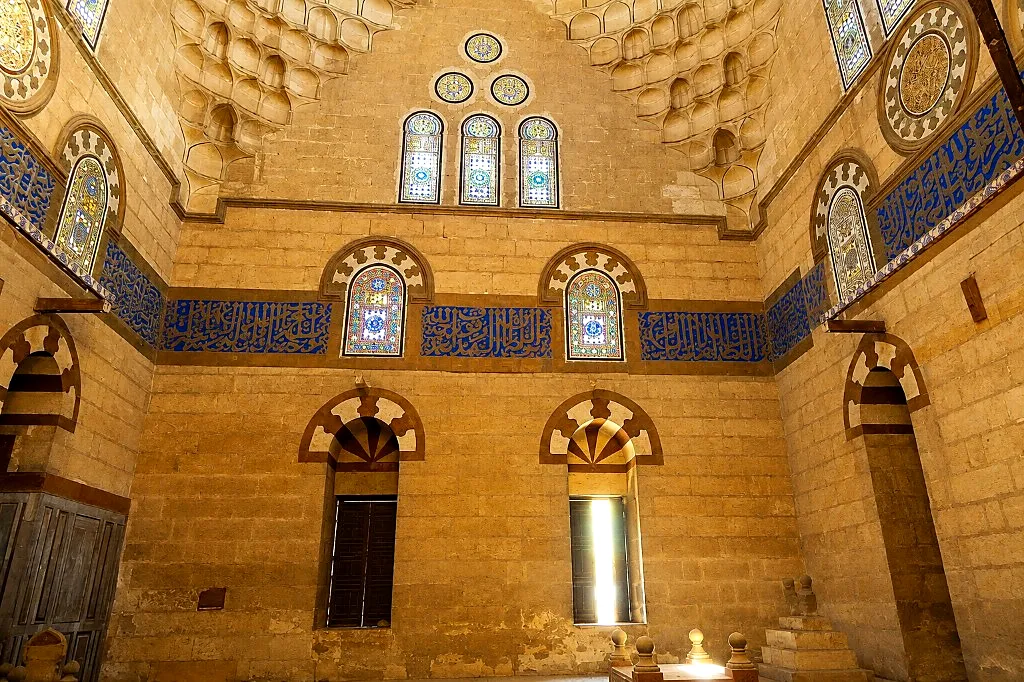 Interior of a Mamluk funerary mosque with blue calligraphy band, stained-glass windows, and stone muqarnas