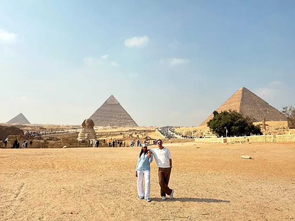 Couple standing on open Giza sand with the Great Sphinx and all three pyramids visible across a wide desert view