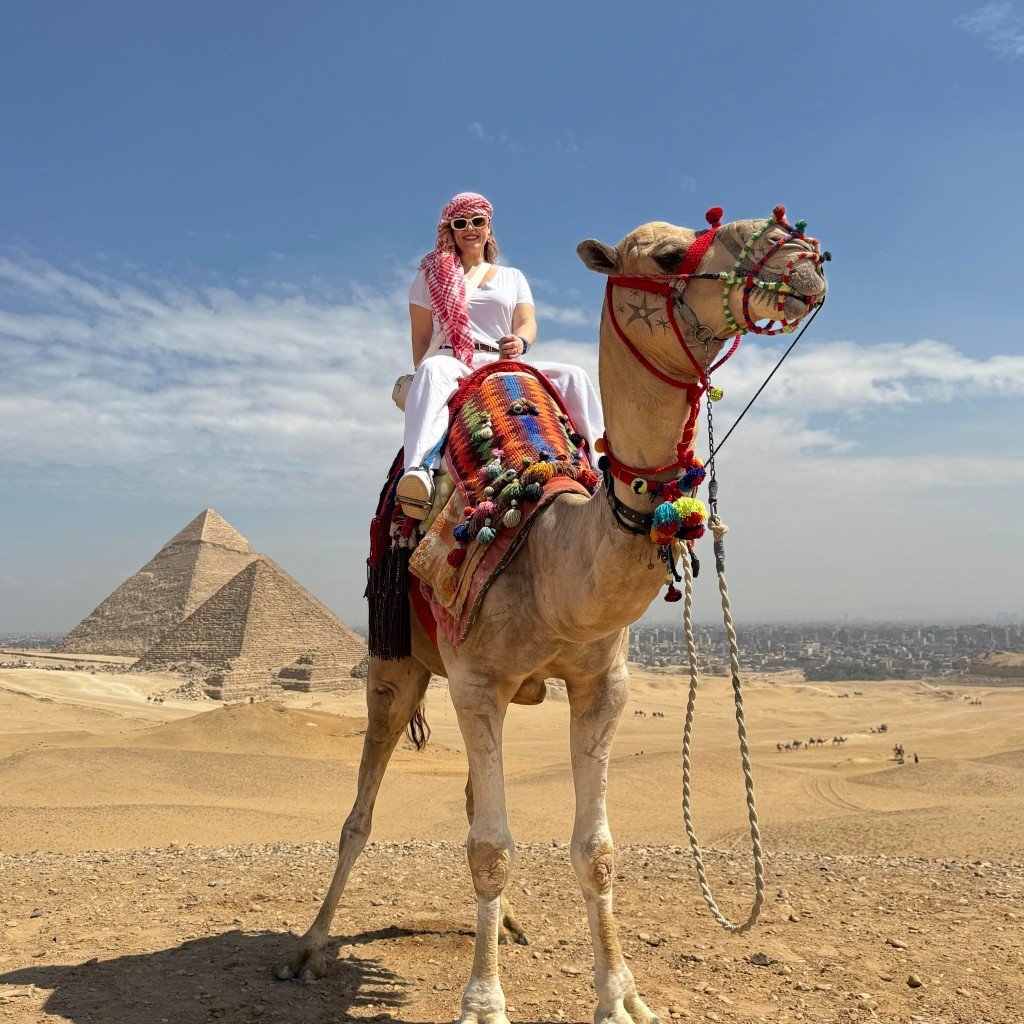 Traveler riding a decorated camel with the Pyramids of Giza behind her on the desert plateau