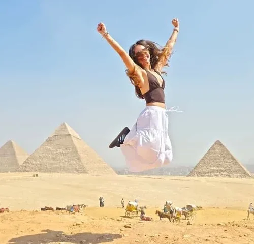 Traveler jumping with arms raised in celebration with three Giza pyramids across the desert behind her