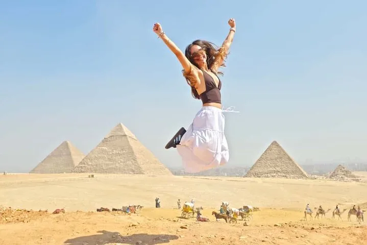 Traveler jumping with arms raised in celebration with three Giza pyramids across the desert behind her