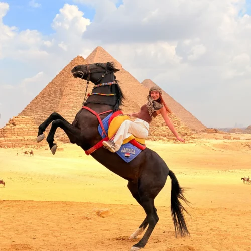 Traveler smiling on a rearing black horse with the Great Pyramid of Giza behind her under a bright blue sky