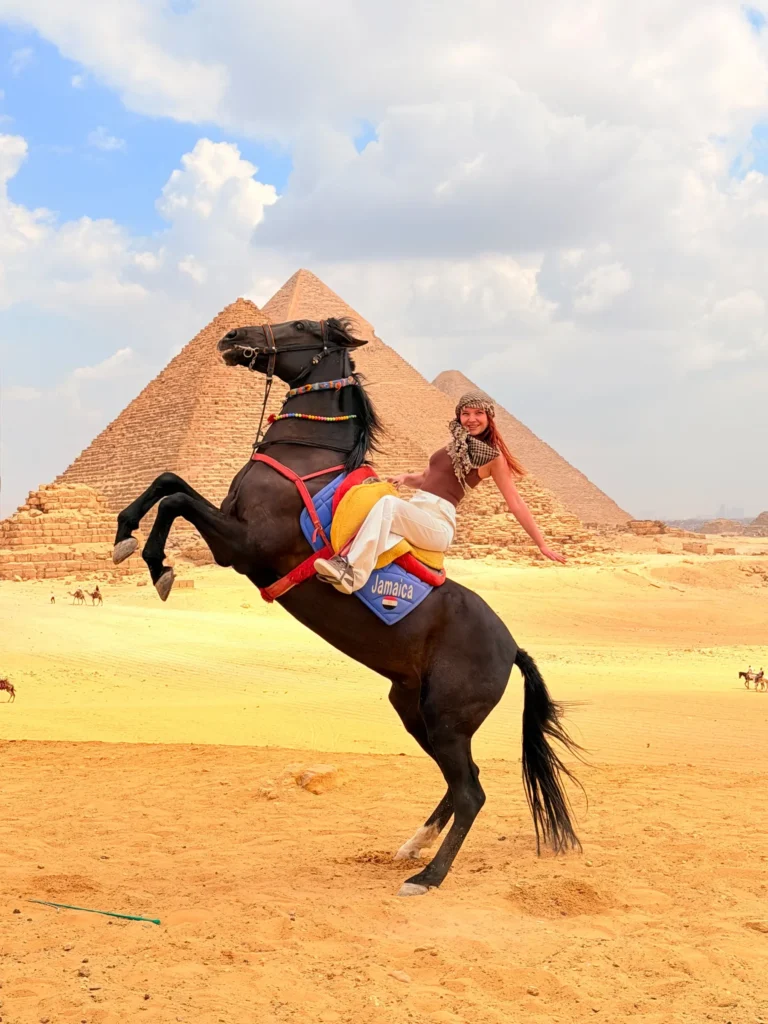 Traveler smiling on a rearing black horse with the Great Pyramid of Giza behind her under a bright blue sky