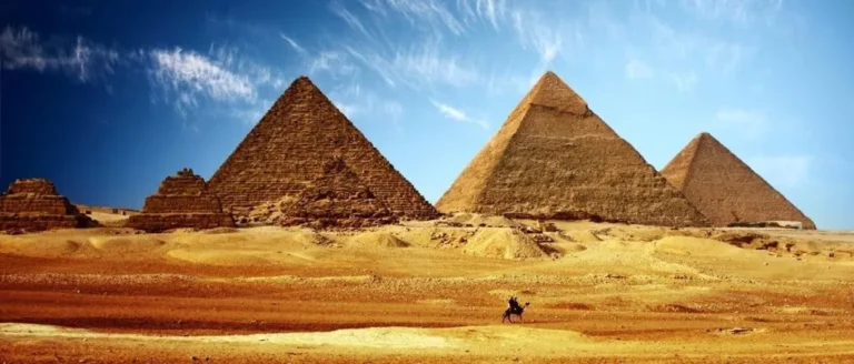 Three Giza pyramids on the desert plateau under a blue sky, with a camel and rider in the foreground sand.