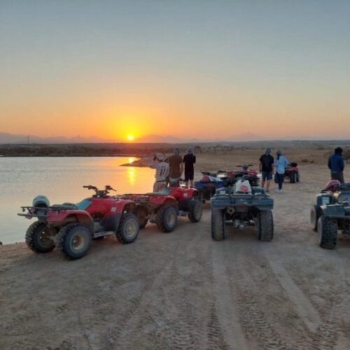 Travelers with red and black quad bikes on desert sand beside water at golden sunset near Hurghada