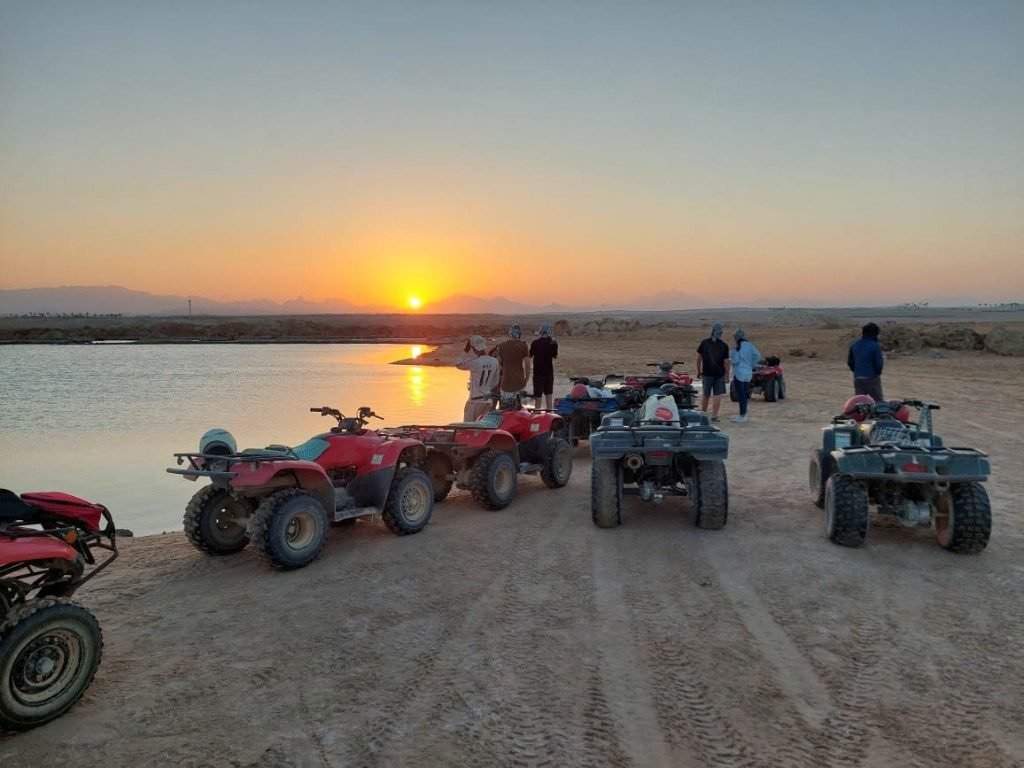 Travelers with red and black quad bikes on desert sand beside water at golden sunset near Hurghada