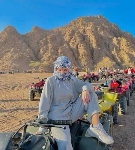 Traveler in a blue checkered shemagh sitting on a green quad bike with a line of ATVs and desert mountains behind