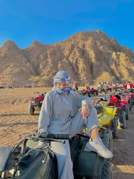 Traveler in a blue checkered shemagh sitting on a green quad bike with a line of ATVs and desert mountains behind