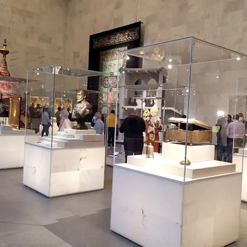 Illuminated display cases of ornate chests and textiles with a ceremonial mahmal tent in an NMEC gallery