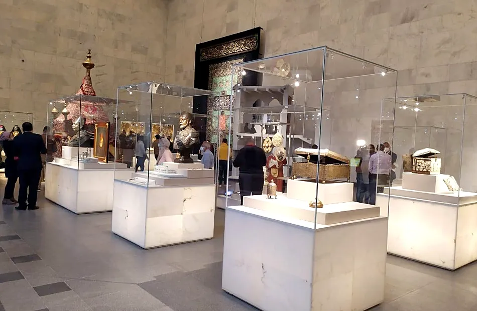 Illuminated display cases of ornate chests and textiles with a ceremonial mahmal tent in an NMEC gallery