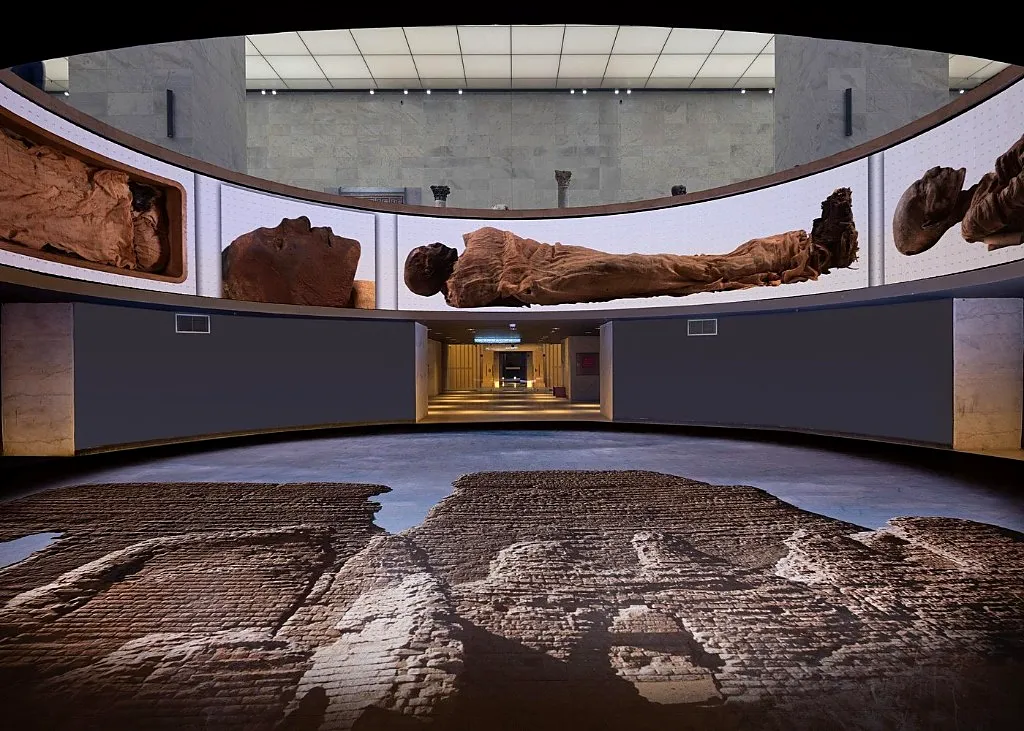 Curved interpretive wall in NMEC showing large photographs of royal mummies above a reconstructed excavation floor