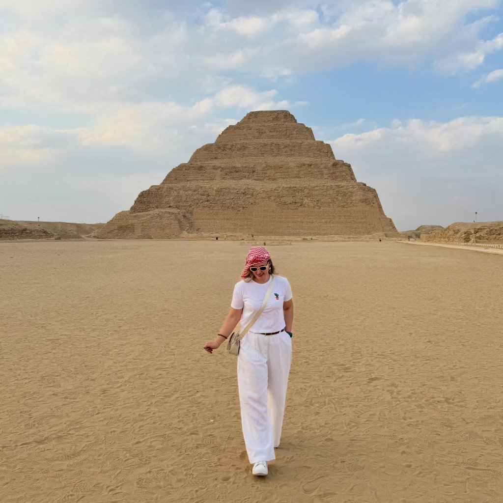 Traveler in a red-and-white keffiyeh walking toward the camera with the Step Pyramid of Djoser at Saqqara behind her