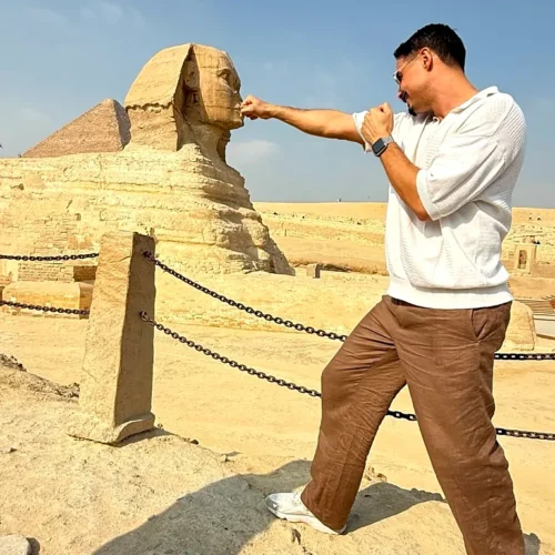 Traveler in a white polo reaching a fist toward the chin of the Great Sphinx in a forced-perspective photo at Giza