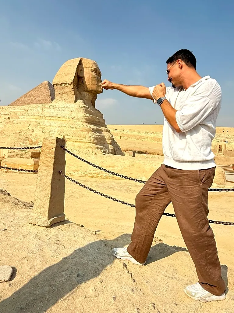 Traveler in a white polo reaching a fist toward the chin of the Great Sphinx in a forced-perspective photo at Giza