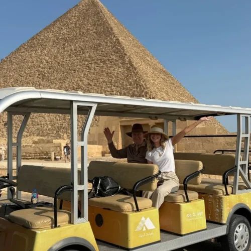 A couple in sun hats waving from a yellow official golf cart in front of a Great Pyramid at Giza