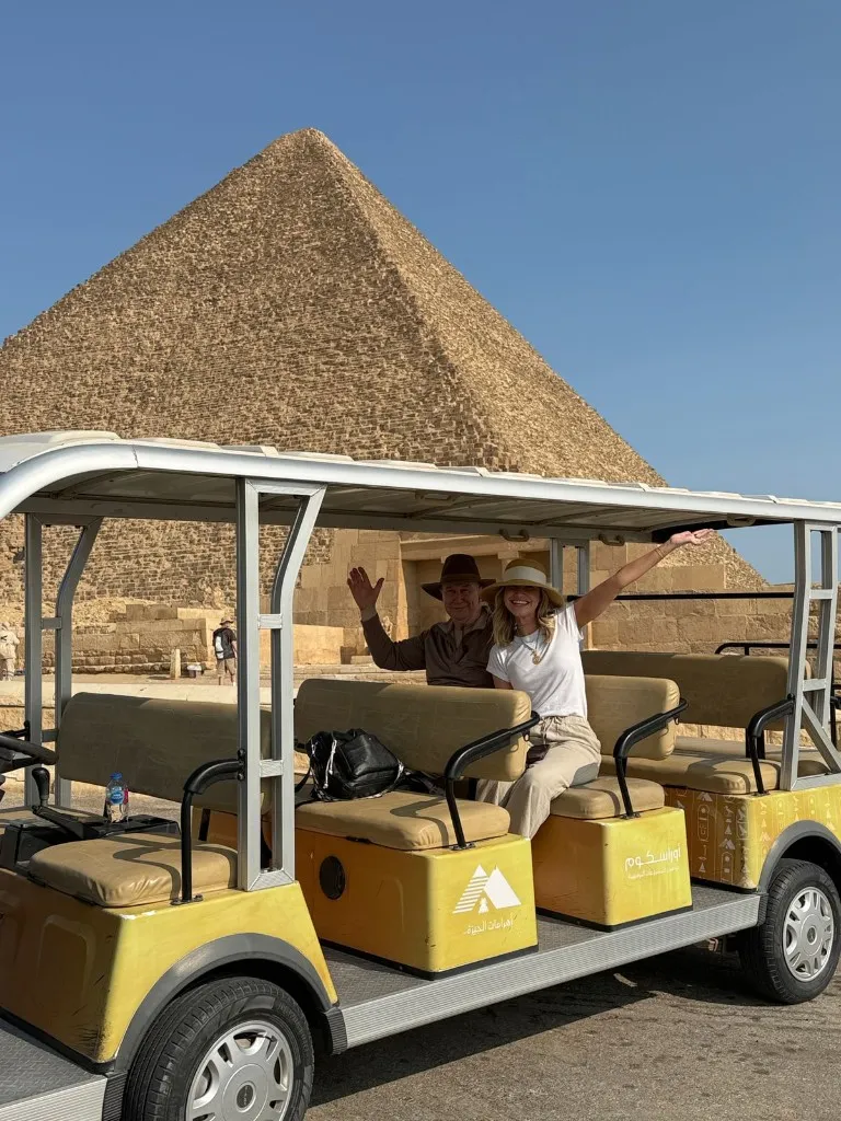 A couple in sun hats waving from a yellow official golf cart in front of a Great Pyramid at Giza