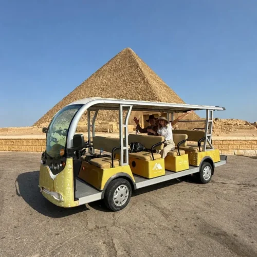 Two travelers waving from a yellow golf cart parked on cracked asphalt in front of a Giza pyramid