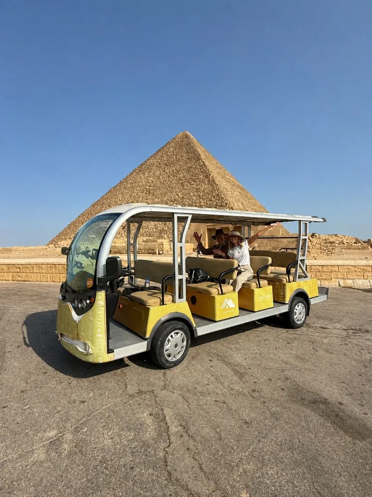 Two travelers waving from a yellow golf cart parked on cracked asphalt in front of a Giza pyramid