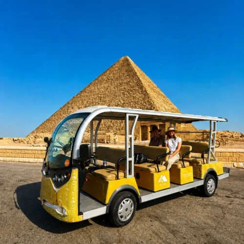 Front three-quarter view of a yellow official Giza golf cart on asphalt, with two travelers and a Great Pyramid behind