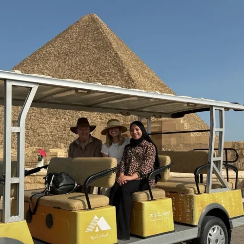 Three travelers seated in a yellow official Giza Pyramids golf cart with a Great Pyramid and clear blue sky behind them