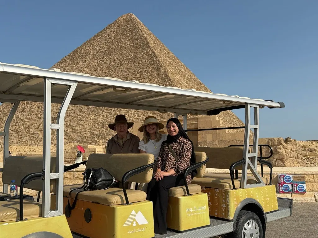 Three travelers seated in a yellow official Giza Pyramids golf cart with a Great Pyramid and clear blue sky behind them
