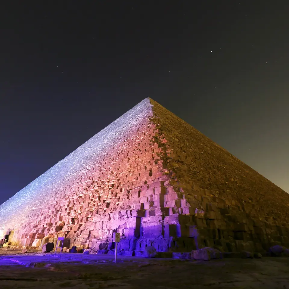A Giza pyramid at night, lit blue near the base and rose higher up the stone.