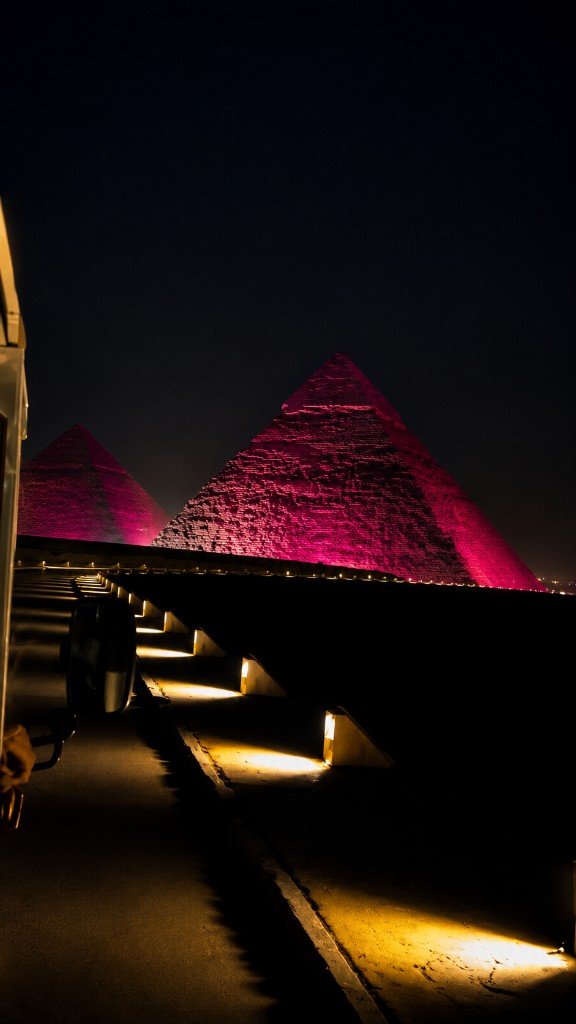 Two Giza pyramids lit deep pink at night, seen past the side of a vehicle.
