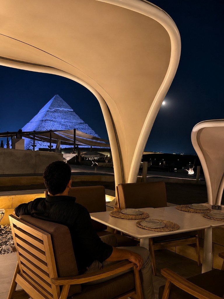 A man sits at a set table under a canopy, looking toward a blue-lit pyramid.