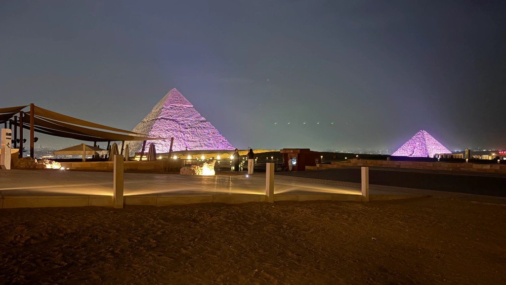 Two Giza pyramids lit purple at night across an open plaza.