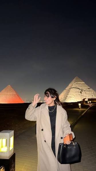 A woman in a long beige coat waves on the Giza plateau walkway, two pyramids lit behind her.
